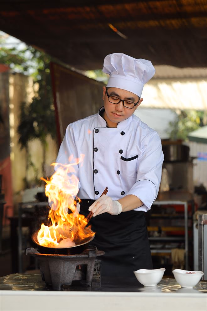 Nguyễn Tiến Hải trong bộ đồ bếp trưởng, nướng gừng và hành trên ngọn lửa lớn giữa bếp mở.