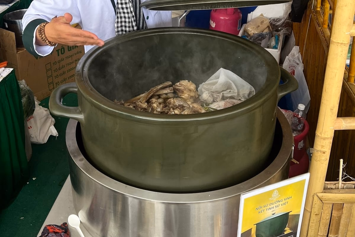Chiếc nồi sứ 100 lít màu xanh rêu đã mở nắp, bên trong là xương bò đang ninh, hơi nước bốc lên; một bàn tay đầu bếp đưa qua miệng nồi.