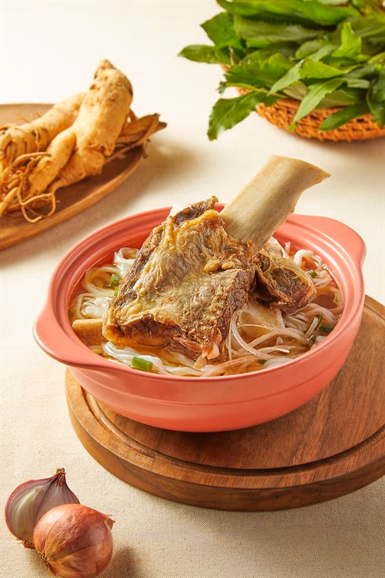 Tảng sườn bò hầm nguyên xương tủy đặt trên bánh phở trong niêu sứ đỏ, quanh là nhân sâm, húng quế và hành tím.